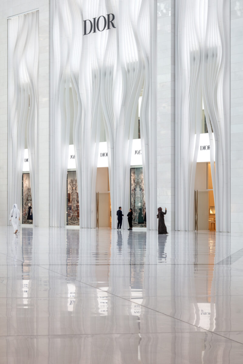 Tall, white Dior store facade with wavy vertical designs; several people walk on a glossy marble floor, some wearing traditional Middle Eastern clothing.