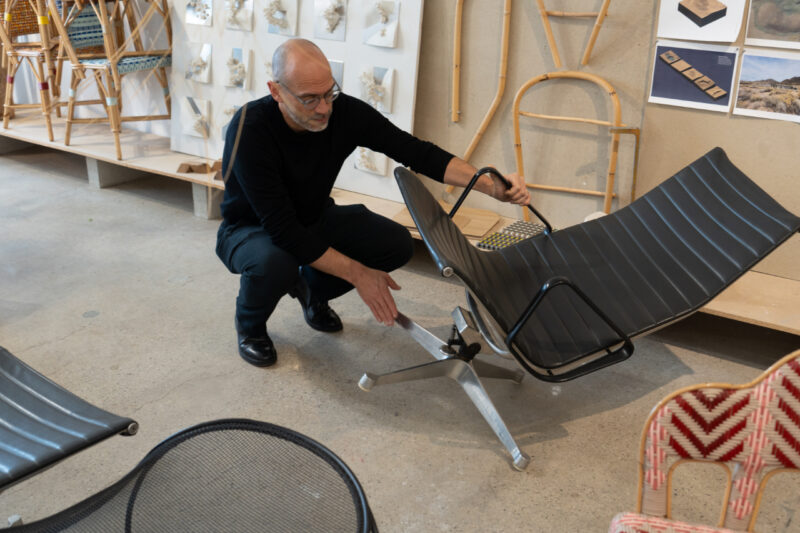 A man wearing glasses and black clothes examines a modern black reclining chair with a metal base in a design studio filled with chairs and sketches.
