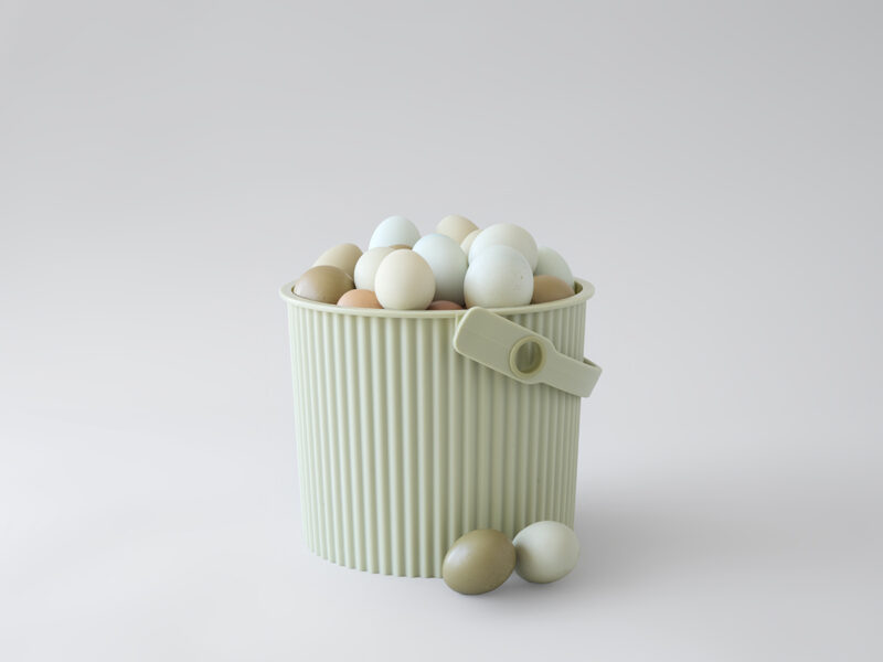 A ribbed, pale green bucket—evoking Martha Stewart’s signature style—filled with multicolored pastel eggs sits on a white surface, with a few eggs and a clothespin resting nearby.