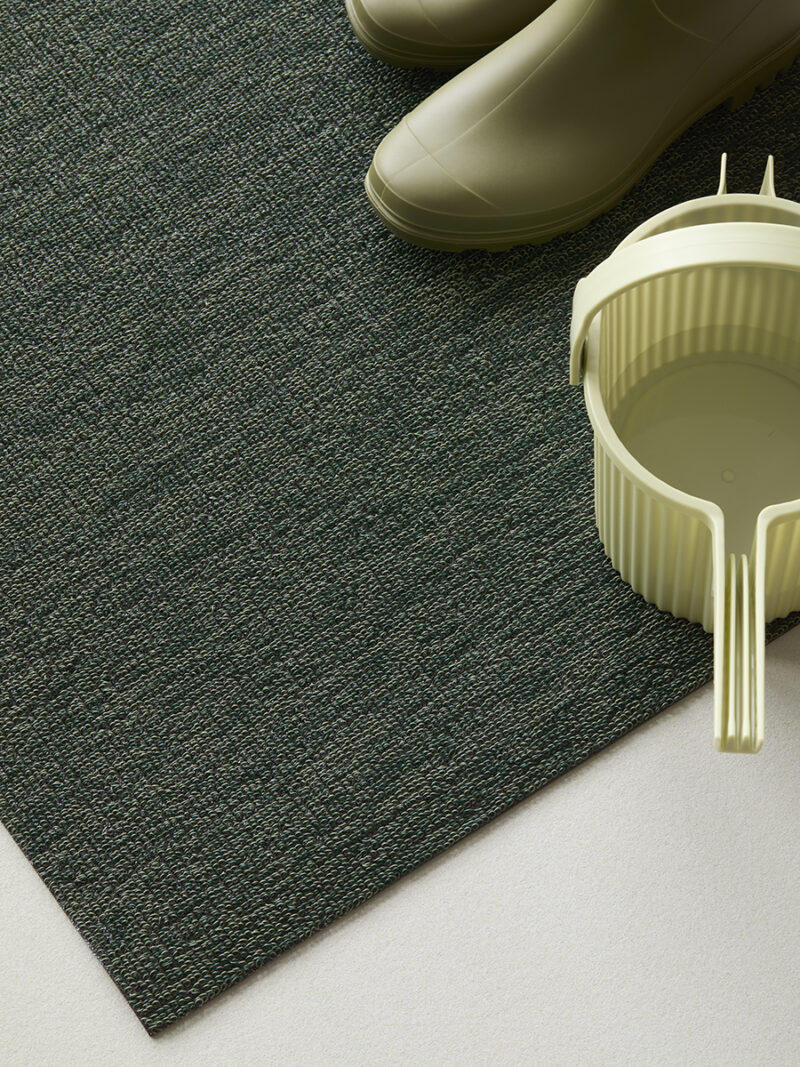 Green rubber boots and a light green plastic bucket are placed on a textured dark green rug, which sits on a light-colored floor—an arrangement reminiscent of Martha Stewart’s classic style, with a touch of Hachiman practicality.