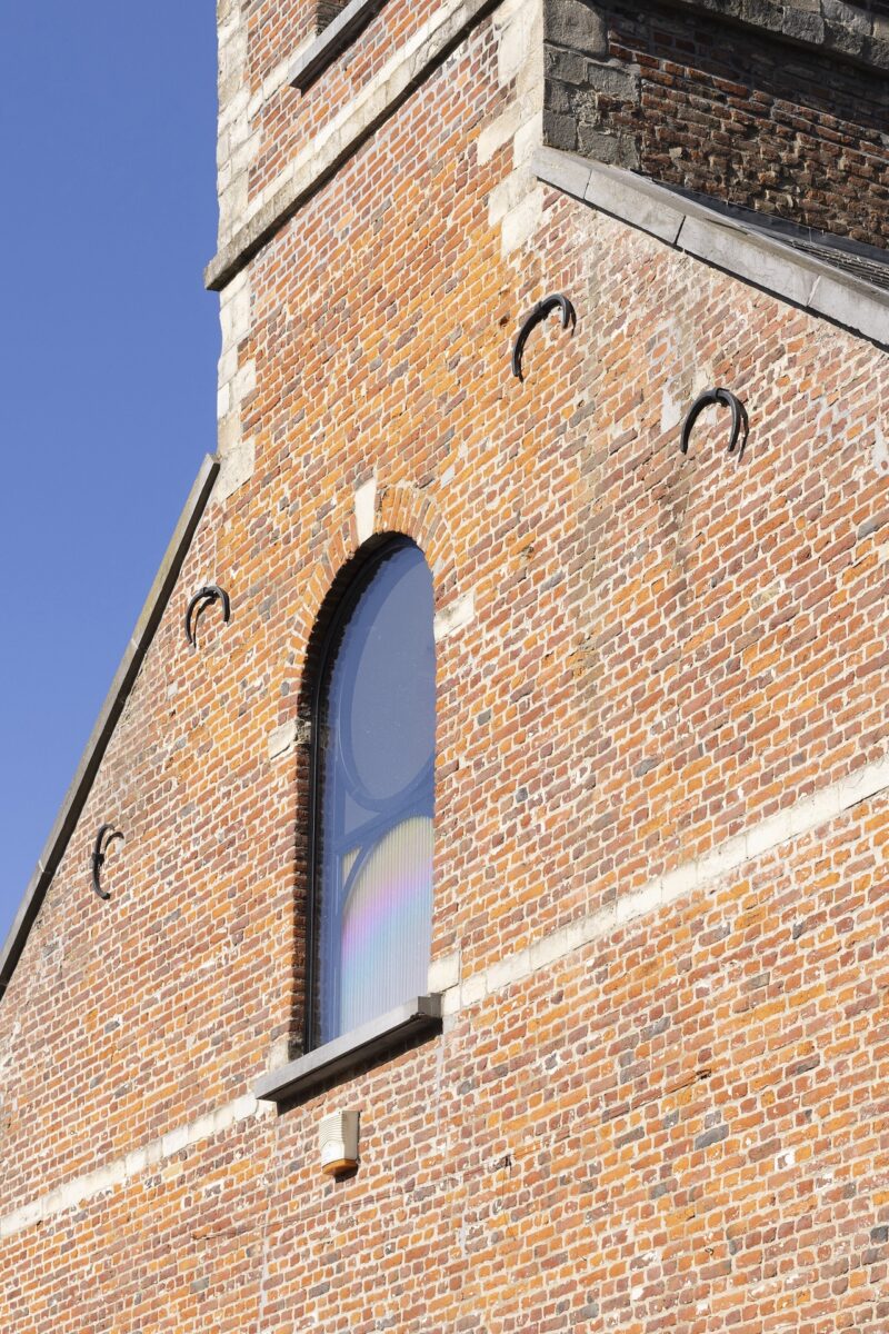 A brick building with an arched window reflects a rainbow pattern. Four metal hooks protrude from the wall, and the sky is clear and blue.
