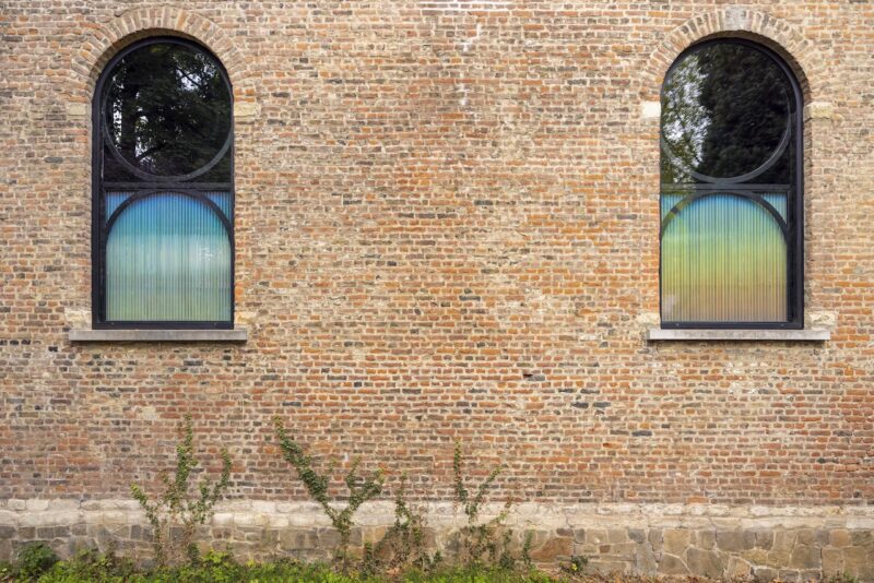 Two arched windows with gradient-colored glass set in a brick wall; greenery grows at the base of the wall.