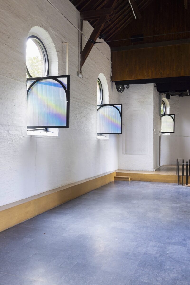 A large room with white brick walls, arched windows, and two square frames with translucent panels displaying rainbows, mounted in front of the windows.