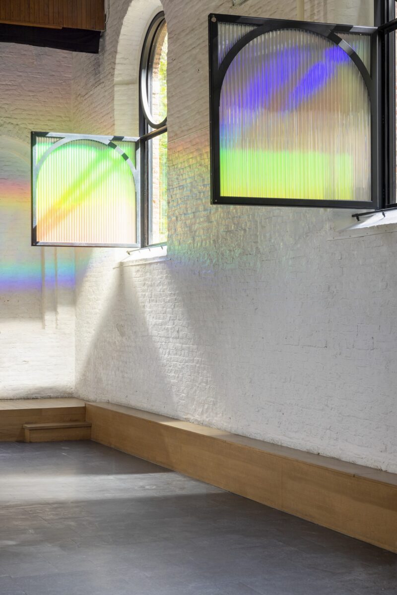 Two arched windows with translucent panels cast rainbow-colored light patterns onto white brick walls and a gray floor in a minimalist room.