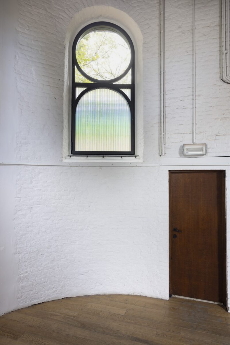 A tall arched window with frosted and clear glass is set above a closed wooden door in a white brick wall, with wooden flooring below.
