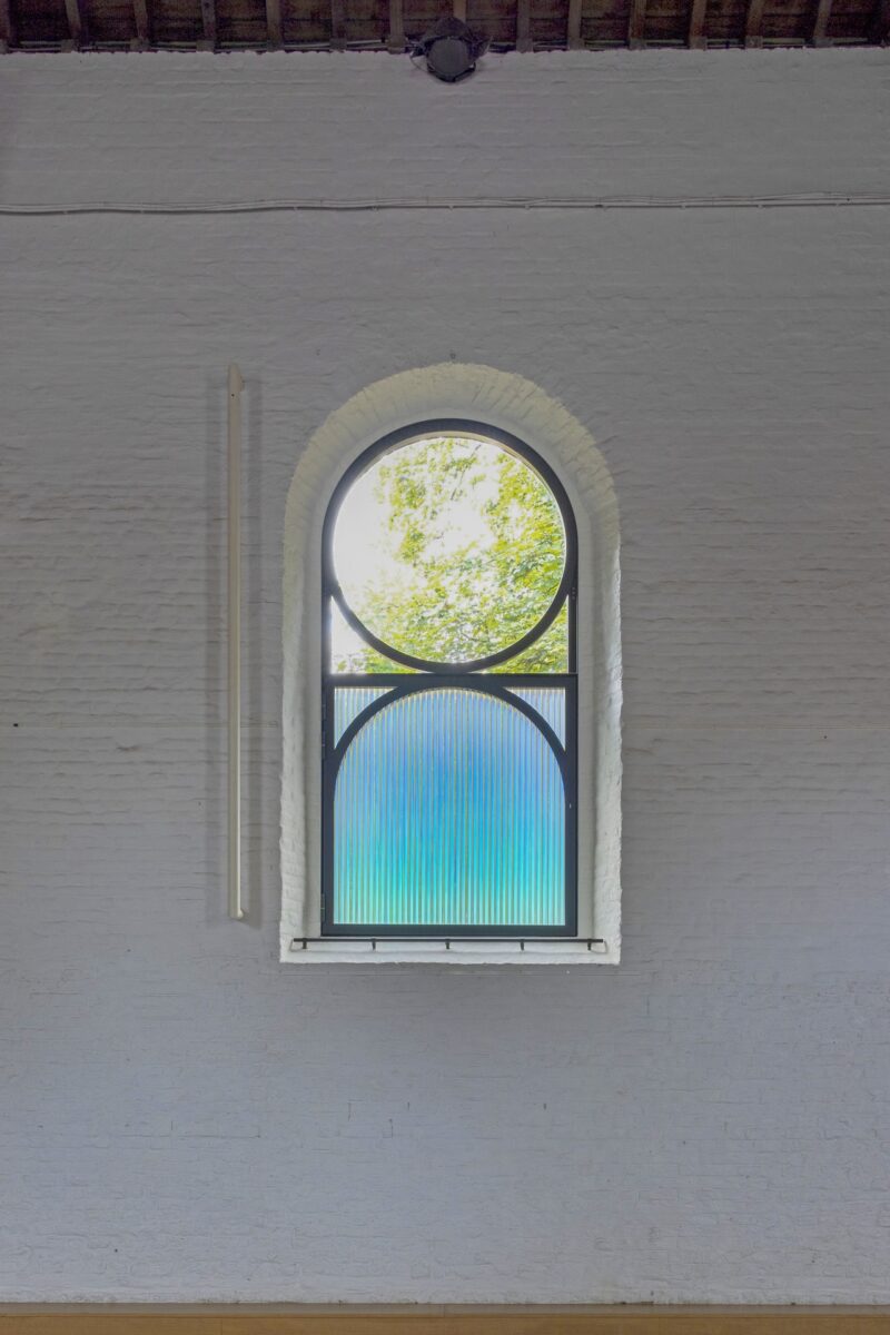 Arched window with circular divided glass; upper part is clear showing green trees outside, lower part is frosted blue, set in a white brick wall.