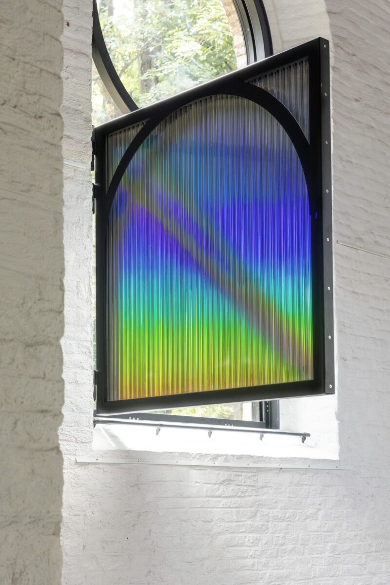 A translucent, ribbed window with a black frame displays a rainbow gradient, allowing light to enter a white brick-walled room. Trees are visible outside through an arched window above.