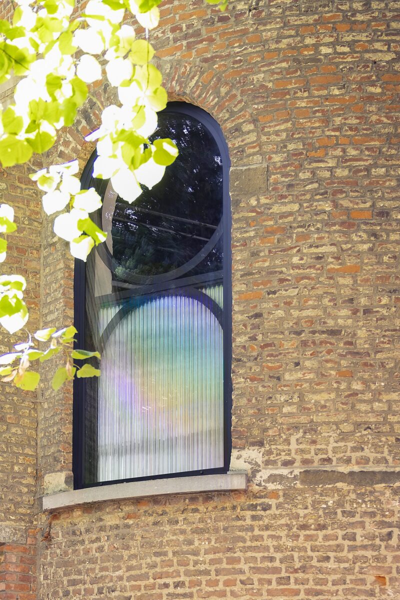 An arched window with a corrugated, partially rainbow-tinted glass pane set in a brick wall, with sunlit green leaves in the upper left foreground.