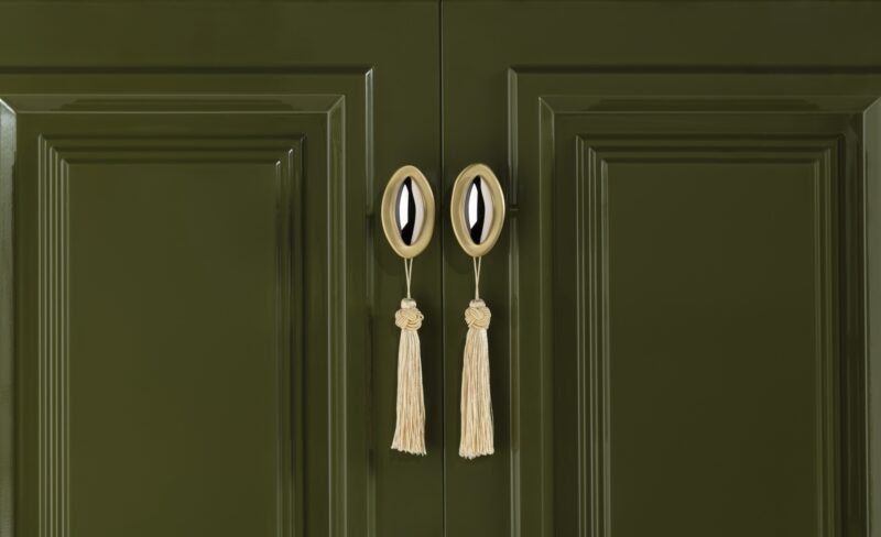 Glossy olive green cabinet doors with gold oval handles, each decorated with a cream-colored tassel tied in a knot, shown in a symmetrical close-up.