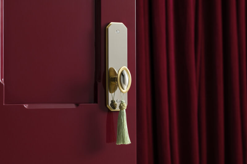 Gold door handle with a green tassel keychain on a glossy burgundy door, next to burgundy curtains.
