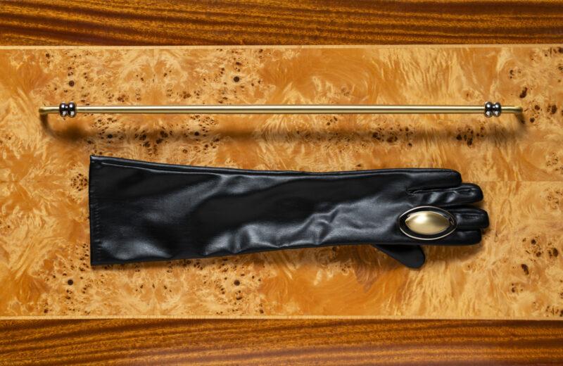 A black leather glove with a large gold button lies flat below a horizontal gold rod on a polished wooden surface.
