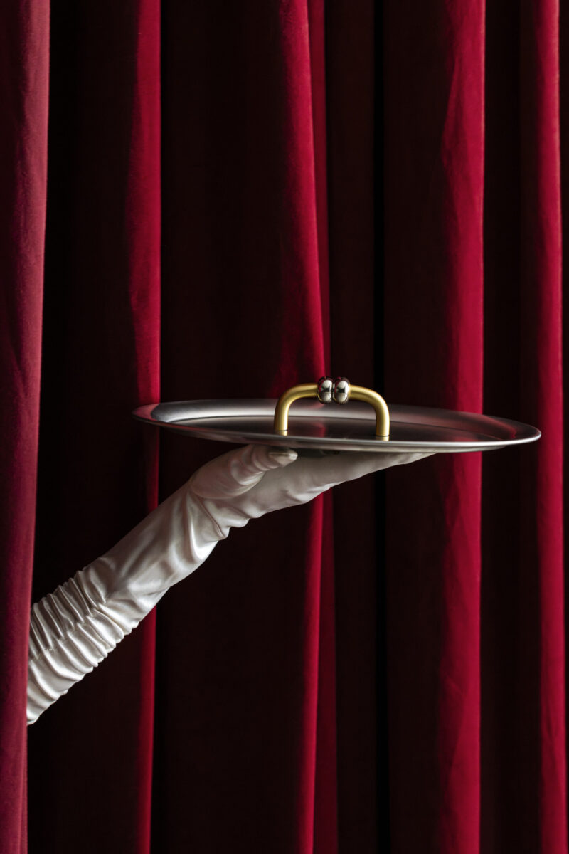 A gloved hand holds a silver tray with a gold handle and two rings on top, set against deep red curtains.