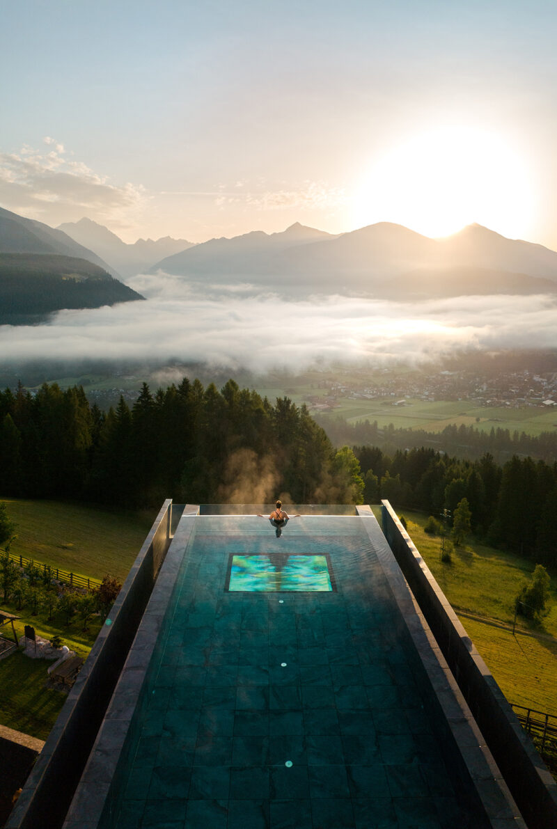 Person sits at the edge of an elevated pool, part of the Hotel Hubertus spa expansion, overlooking a misty valley with mountains and a sunrise in the background.