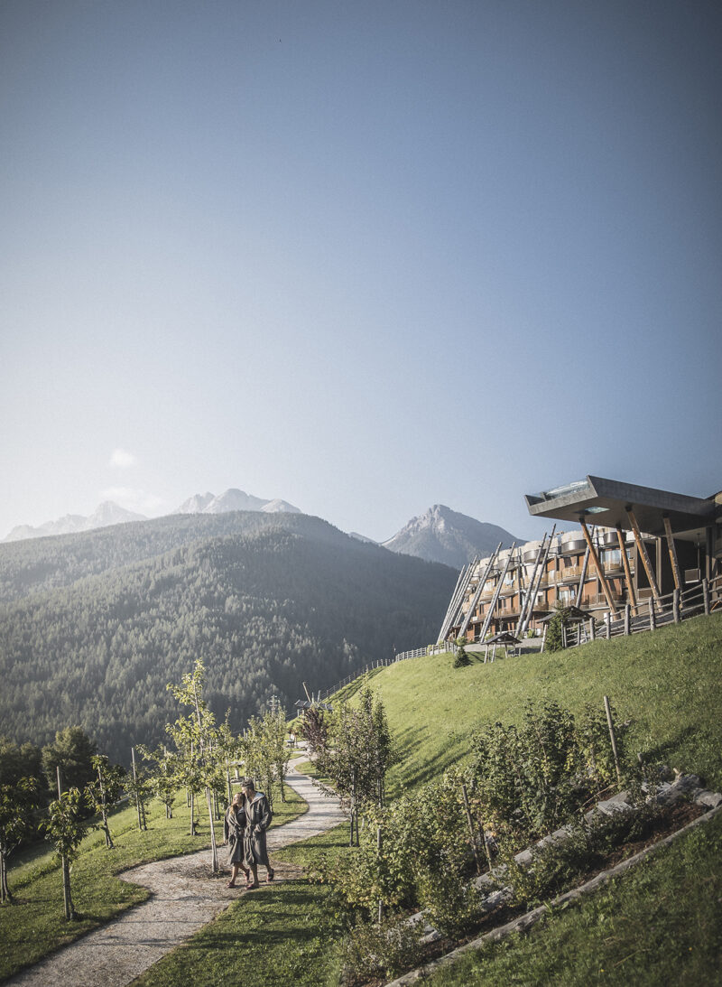 Two people walk along a winding path on a grassy hillside, with the modern wooden buildings of the Hotel Hubertus spa expansion and mountain peaks visible in the background under a clear sky.