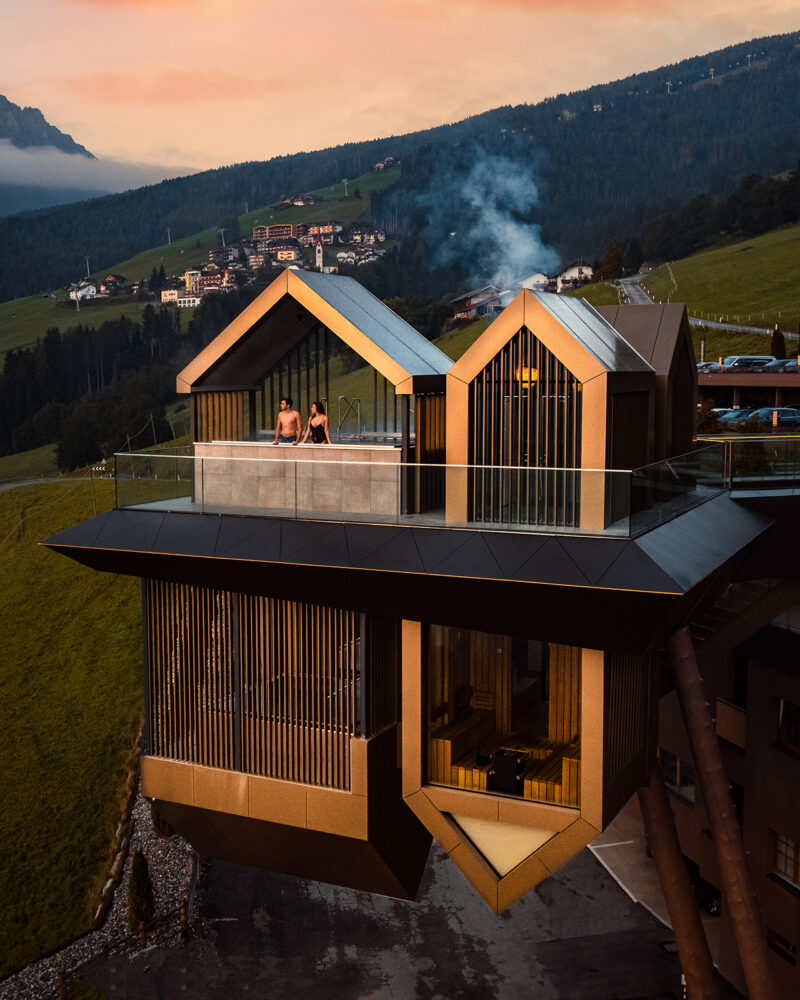 Modern building with angular architecture extends over a hillside; two people sit in a rooftop hot tub, part of the Hotel Hubertus spa expansion, overlooking a scenic rural landscape with mountains and scattered houses at sunset.