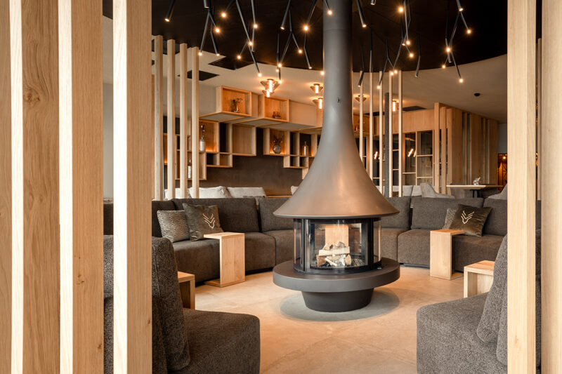 Modern lounge with a central suspended fireplace, gray sectional sofas, wooden accents, open shelving, and overhead pendant lights—an inviting space reminiscent of the sophisticated elegance found in the Hotel Hubertus spa expansion.