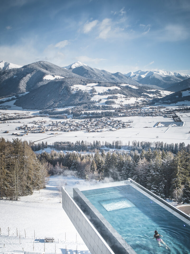 An outdoor pool, part of the Hotel Hubertus spa expansion, extends over a snowy landscape with mountains and a village in the background; one person swims in the pool under a clear sky.