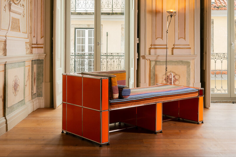 Modern orange modular bench by Armando Cabral with a striped cushion and pillows, set in an ornate room featuring classic wooden floors, tall windows, a floor lamp, and sophisticated USM accents.