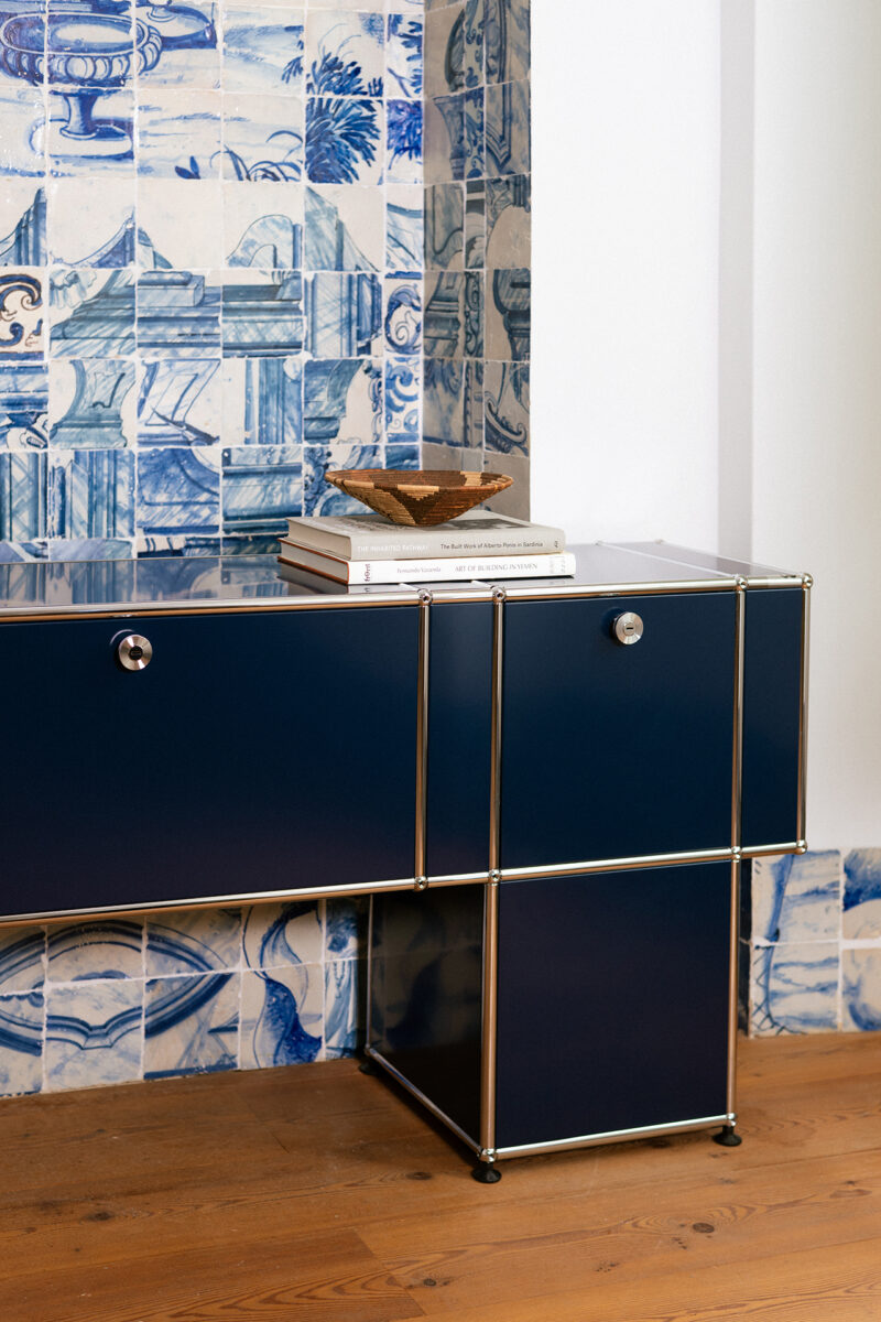 A blue modular cabinet by USM with sleek chrome edges stands on a wooden floor beside blue and white patterned wall tiles; books and a decorative object, curated in the style of Armando Cabral, rest on top.