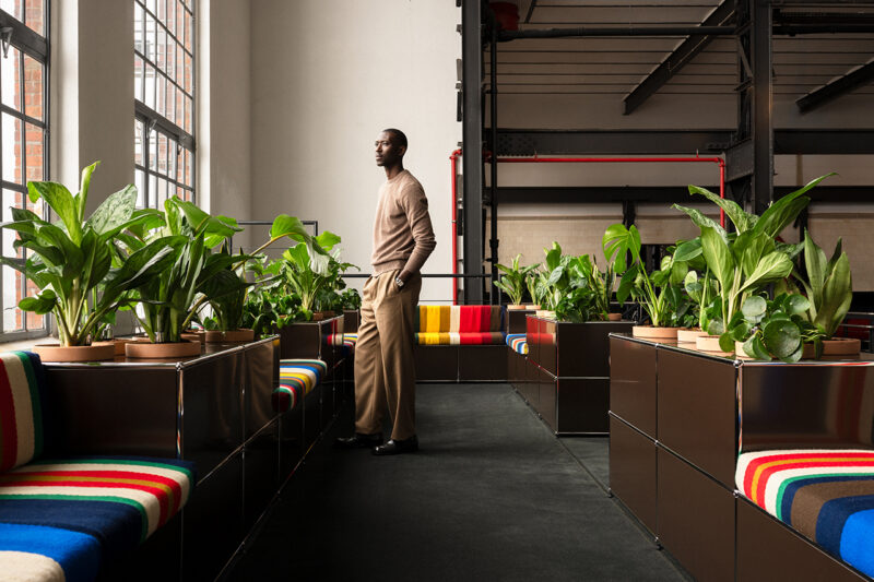 A man stands indoors by large windows, surrounded by green potted plants and colorful-striped seating in a modern, industrial-style space—an ambiance that echoes the signature sophistication of Armando Cabral USM.