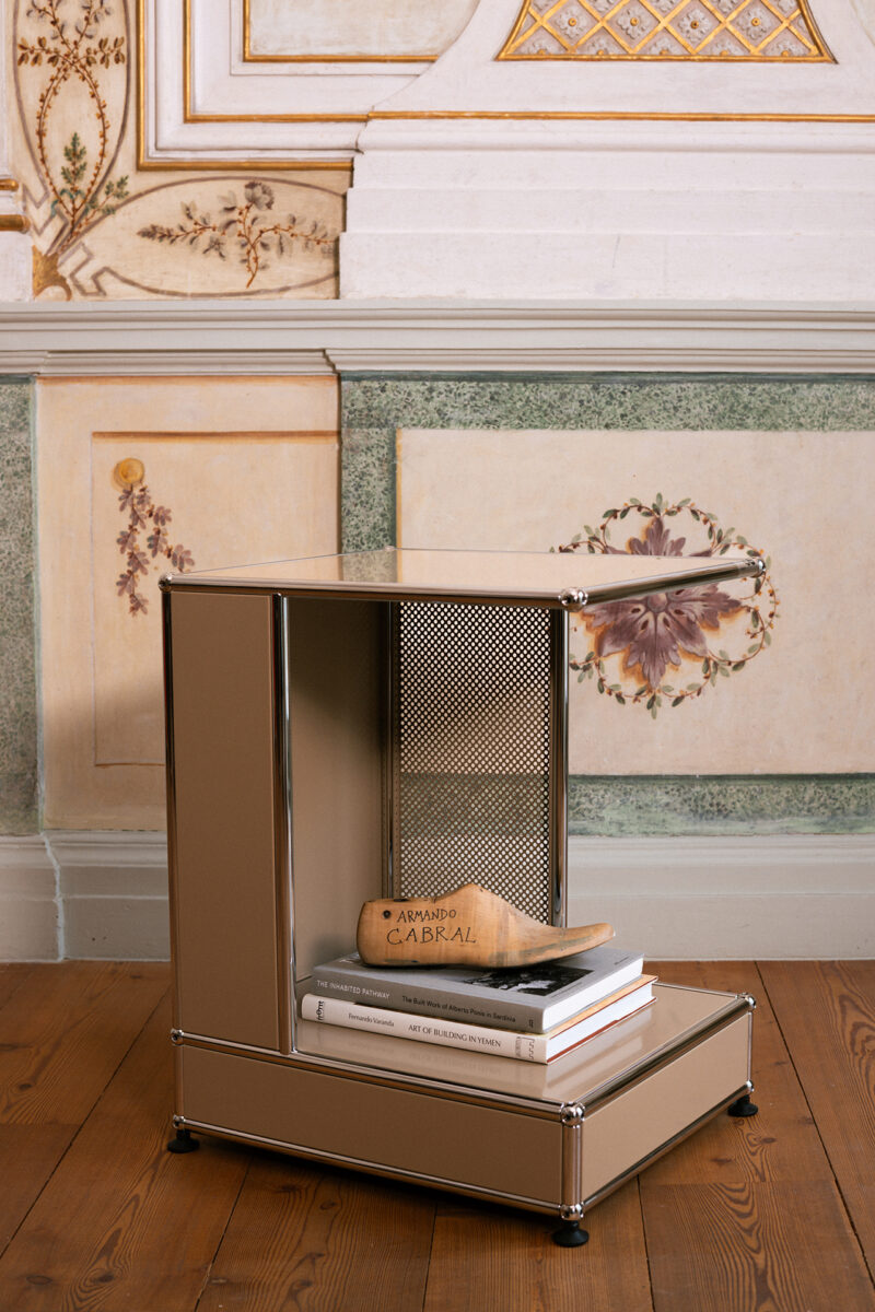 A small beige USM metal side table holds a stack of books and a wooden shoe form, set against an ornate, painted wall—a subtle nod to the refined aesthetics admired by Armando Cabral.