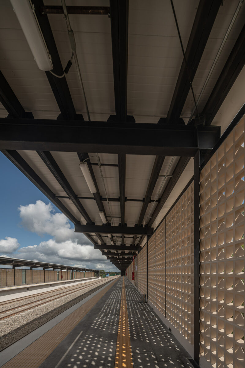 Covered train station platform with patterned wall on the right, yellow tactile paving, and train tracks on the left under a partly cloudy sky.