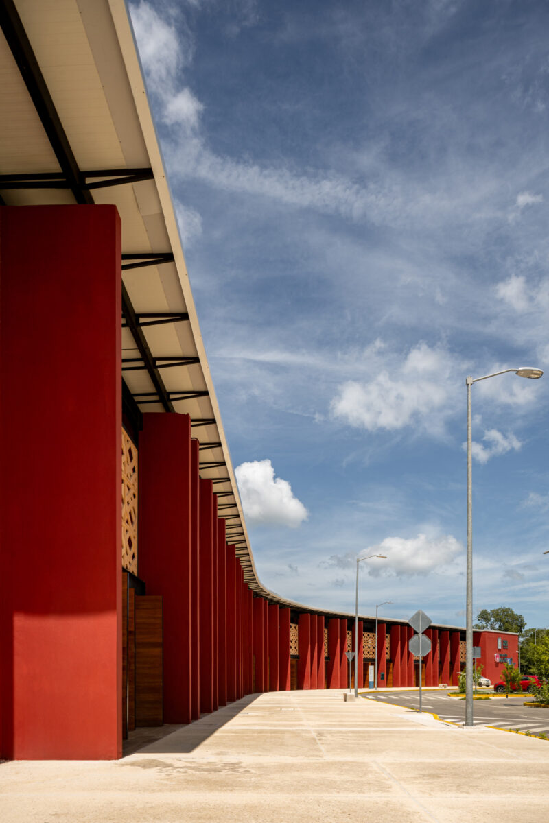A modern building with tall red columns and decorative panels stands under a partly cloudy blue sky, with street lamps and a sidewalk visible.