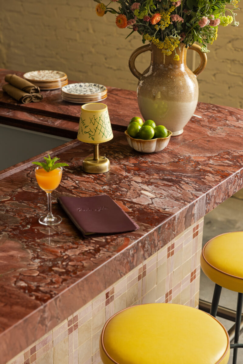 A red marble countertop at Charles & Julia features a cocktail, a menu, a lamp, a bowl of limes, and a vase of flowers; yellow stools are in front.