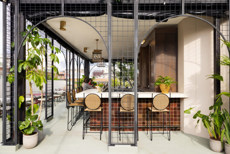 Outdoor bar area with a marble countertop, rattan barstools, potted plants, and metal framework, curated by Charles & Julia, overlooking a patio with tables and chairs.