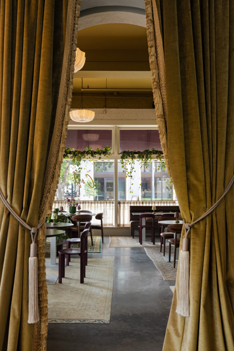 Large gold curtains frame the view into Charles & Julia, a modern restaurant with wooden tables, purple chairs, hanging plants, and large windows letting in natural light.