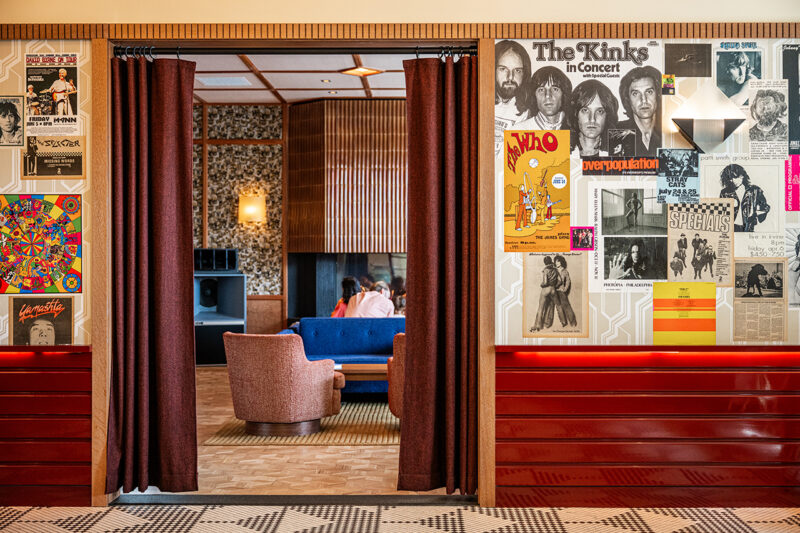 A lounge area at Fishtown Urby is seen through an open curtain, with vintage rock posters on the wall and retro furniture including a pink chair and blue sofa.