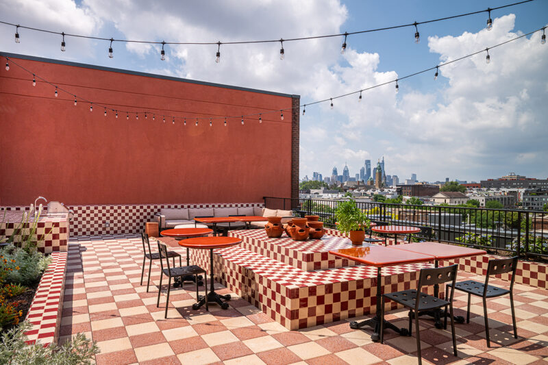 Rooftop patio at Fishtown Urby with red and white checkered tile, tables, chairs, potted plants, string lights overhead, and a city skyline in the background under a partly cloudy sky.