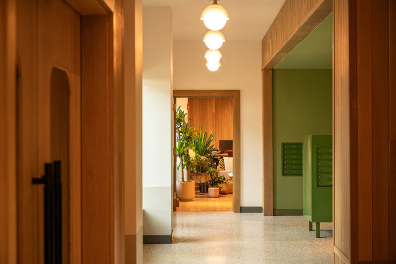 A hallway at Fishtown Urby, with wooden doors, green mailboxes, and hanging lights, leads to a room filled with potted plants and wooden furniture.