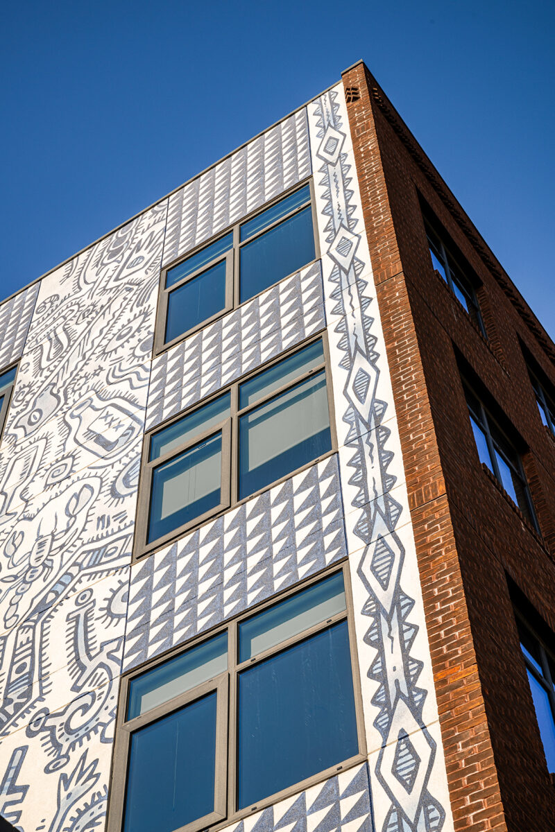 Corner of a brick building with patterned mural art and geometric tile designs under a clear blue sky, capturing the vibrant aesthetic of Fishtown Urby.