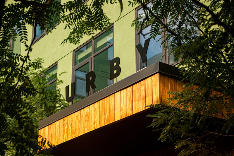 A modern building with green walls and large windows displays the black letters "URBY" on a wooden ledge, partially obscured by tree branches—an inviting sight at Fishtown Urby.