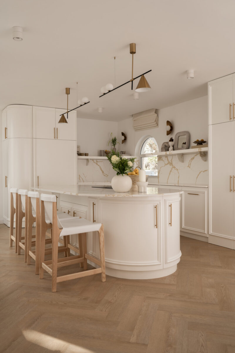 Modern kitchen with white cabinets, marble backsplash, a curved island with four wooden stools, gold fixtures, and neutral décor. Natural light enters through an arched window.
