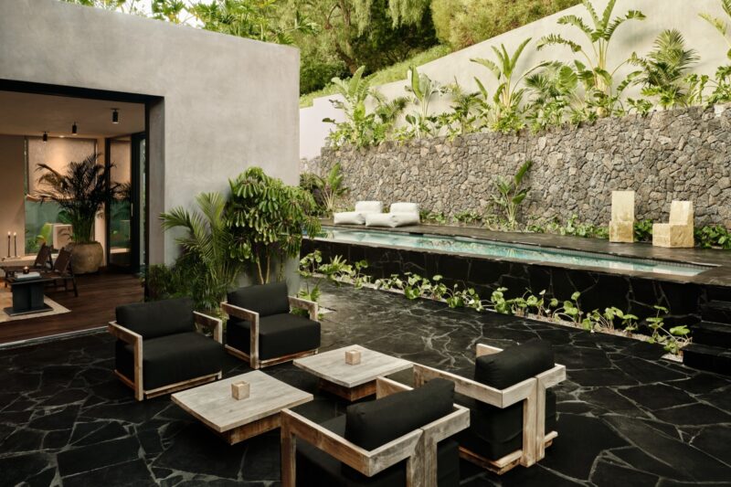 Modern outdoor patio with black stone floor in an elegant Obsidian House style, wooden furniture with black cushions, a small pool, lush plants, and a beautiful stone wall.
