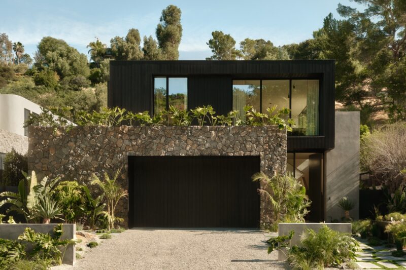 Four woven stools surround a stone fire pit with a small fire, set in the gravel courtyard of Obsidian House, framed by dense tropical plants and a striking stone wall.