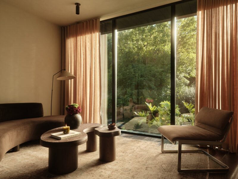 A modern living room at Obsidian House with warm, neutral tones, a curved brown sofa, a wooden coffee table, a sleek chair, and large floor-to-ceiling windows looking out onto a sunlit, leafy garden.