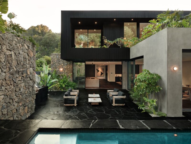Obsidian House: A modern house with large windows, outdoor seating area, striking stone walls, lush plants, and a swimming pool in the foreground.