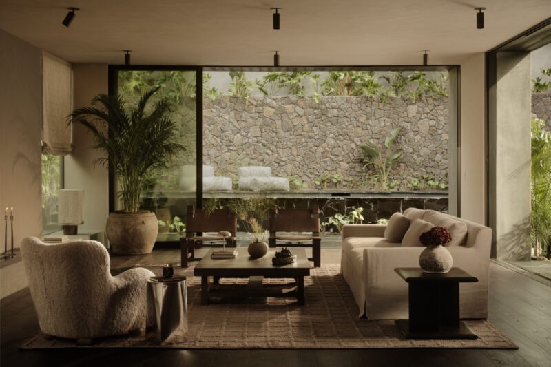 Modern living room with minimalist furniture, large windows, and a view of a stone wall and lush greenery outside evoke the serene elegance of an Obsidian House. Neutral tones and natural light create a calm, cozy atmosphere.