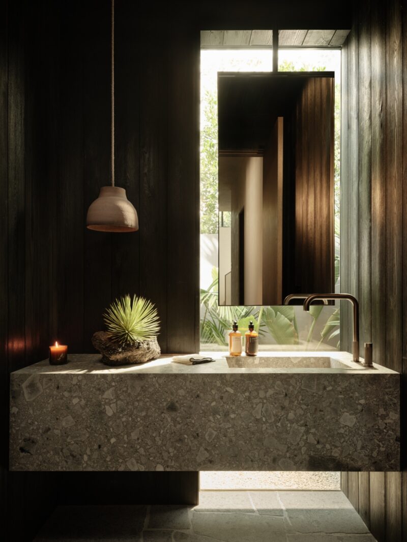 Modern bathroom inside Obsidian House with a stone sink, wall-mounted faucet, decorative plant, candle, toiletries, and a mirror reflecting outdoor greenery. Natural light enters through a tall window.