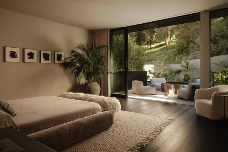 A modern bedroom in Obsidian House features a large bed, potted plant, wall art, and sliding glass doors opening to a patio with outdoor seating and lush greenery.