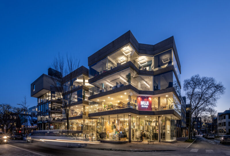 A modern, multi-story building at dusk features large glass windows and illuminated interiors. A red "MUJI" store sign is visible on the ground floor, marking the MUJI Harvard Square location. Trees and street traffic are in the foreground.