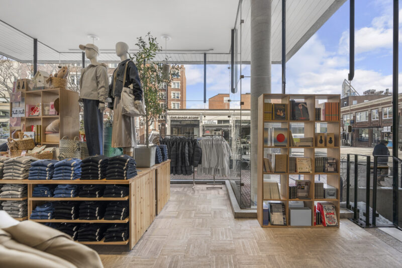 Modern retail store interior with mannequins in casual clothes, shelves of folded jeans, jackets on racks, and a display of books and magazines by large windows showing a city street outside—evoking the minimalist aesthetic found at MUJI Harvard Square.