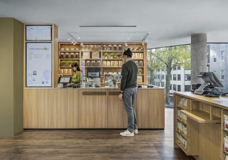 A person stands at a modern coffee shop counter while a barista prepares an order. Shelves behind the counter display coffee bags and plants, evoking the minimalist aesthetic of MUJI Harvard Square. Large windows reveal trees and buildings outside.