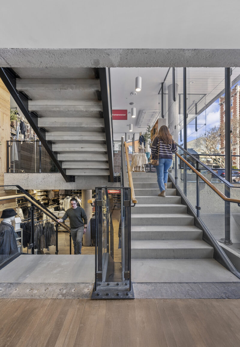 A woman walks up a modern indoor staircase with glass railings at MUJI Harvard Square, while another person descends. The area features large windows and retail displays.