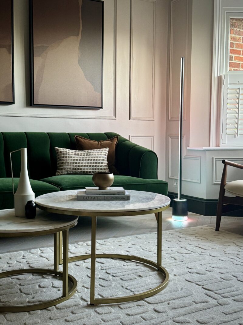A modern living room with a green sofa, round marble coffee tables, decorative vases, wall art, and a tall floor lamp near a window with white shutters features thoughtfully layered lighting to create a warm and inviting atmosphere.