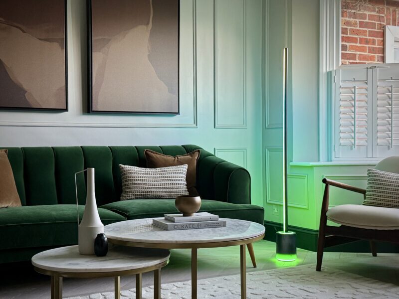 Modern living room with a green velvet sofa, abstract wall art, round coffee tables, decorative vases, layered lighting from a glowing floor lamp, and a window with white shutters.