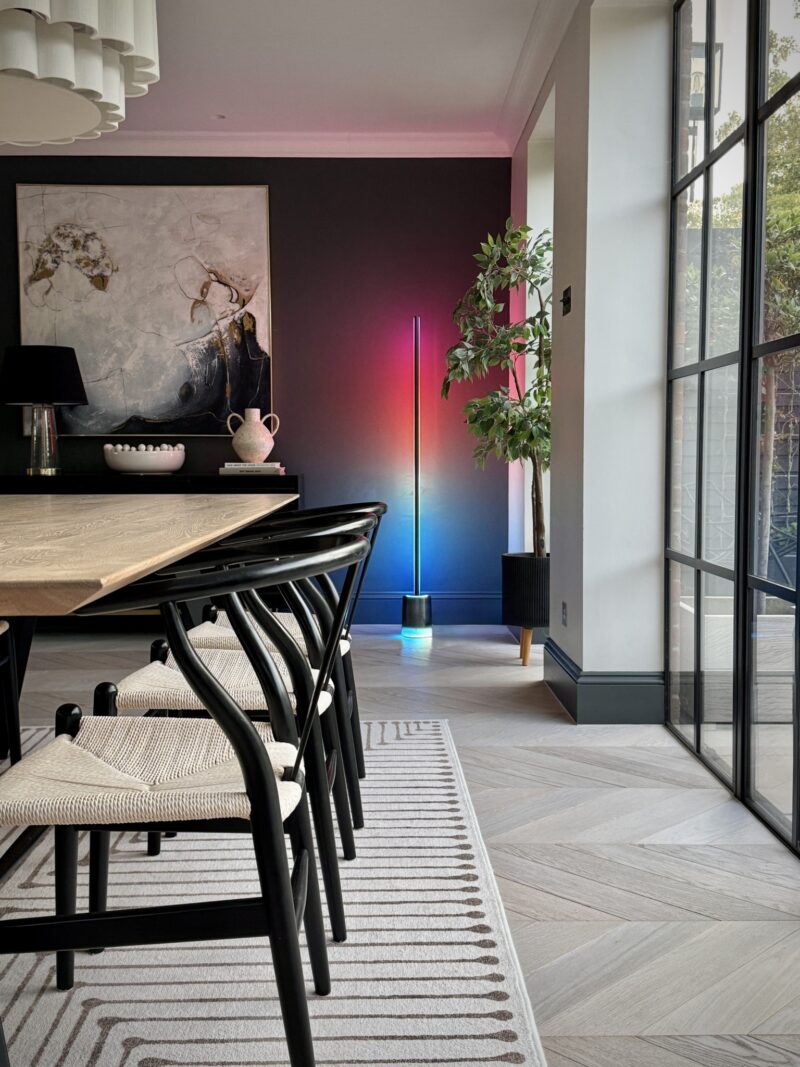 Modern dining room with a wooden table, black chairs, abstract wall art, a large plant, and a colorful floor lamp providing layered lighting against a dark accent wall near large windows.